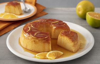 Receita de Pudim de Laranja. Observa-se um prato grande branco contendo o pudim com calda e raspas. Atrás, um prato de sobremesa com uma fatia do pudim e uma colherinha. Ao lado, laranjas decoram a foto.