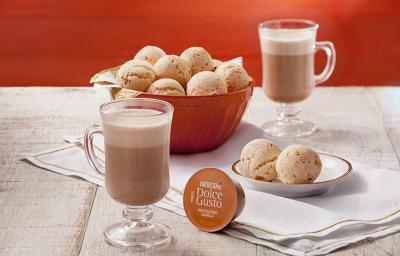 fotografia em tons de laranja e cinza de uma bancada cinza vista de cima, contém um pano branco, um recipiente redondo laranja com vários pães de queijo, duas xicaras transparentes com Mochaccino Canela, um prato redondo com dois pães de queijo.