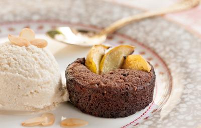 Fotografia em tons de salmão em uma bancada de madeira com uma toalha branca com detalhes salmão, um prato redondo de porcelana desenhado em cinza e rosa com uma bola de sorvete de creme e um mini bolinho de chocolate em cima dele.