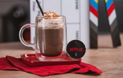 Foto em tons de vermelho e preto da receita de cappuccino netflix servida em um copo largo sobre uma base de porcelana vermelha e um pano vermelho em cima de uma mesa de madeira com uma cápsula de cappuccino netflix ao lado