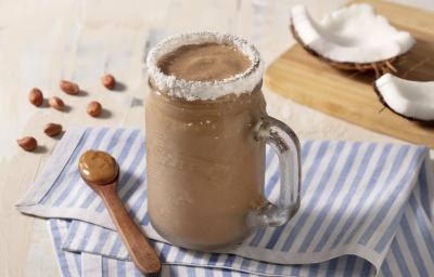 Pano listrado de azul claro e branco, com uma colher de madeira marrom com pasta de amendoim. Uma caneca transparente com o smoothie e borda de coco ralado.