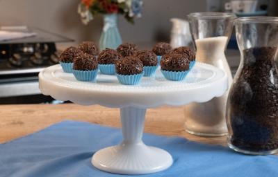 Fotografia de dez forminhas azuis com brigadeiros passados no granulado e apoiados em um enfeite para bolo branco e alto. Ao fundo tem um fogão, um vaso azul com flores, e ao lado do brigadeiro tem duas jarras decorativas de vidro com açúcar e cacau.