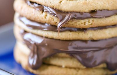 Fotografia de biscoitos empilhados, e entre alguns, um creme de chocolate caindo um pouco nas bordas. Os biscoitos estão em um prato quadrado na cor azul em tom mais escuro, que estão sobre uma mesa na cor marrom escuro.