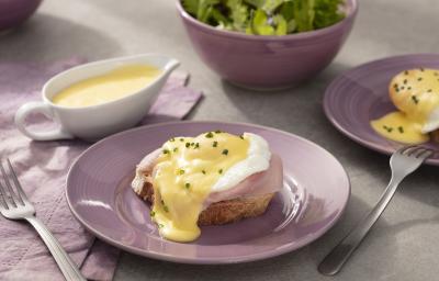 Fotografia em tons de lilás em uma bancada de madeira clara, um paninho lilás, um prato redondo lilás com o ovo benedict em cima dele, com a fatia de pão tostado, presunto, o ovo e cebolinha-verde para decorar. Ao fundo, um bowl com salada verde.