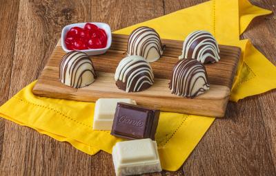 Fotografia em tons de marrom em uma bancada de madeira de cor marrom. Ao centro, um pano amarelo contendo uma tábua de madeira com as trufas e ao lado, há um recipiente contendo cerejas.