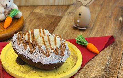 Fotografia em tons de amarelo e vermelho em uma mesa de madeira com um prato amarelo ao centro. Dentro dele existe um ovo de chocolate recheado e coberto com chantilly, doce de leite e banana. Ao fundo tem um coelho e algumas cenouras de pelúcia.