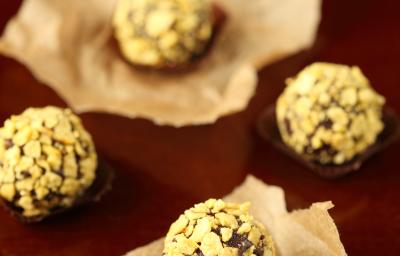 Fotografia de cinco brigadeiros enrolados e passados em nozes, alguns em forminhas, outros sobre um papel manteiga. Ao fundo, forminhas empilhadas, sobre uma mesa marrom em tom escuro.