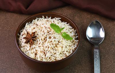 Fotografia em tons escuros de um recipiente fundo na cor marrom com arroz e especiarias e duas folhas de hortelã por cima. Ao lado, uma colher de sopa desenhada, sobre uma mesa marrom em tom escuro.