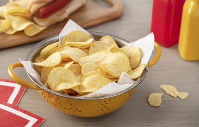 Fotografia em tons de vermelho em uma bancada de madeira clara, um pano vermelho com listra branca, uma tábua de madeira com um cachorro-quente e batatas chips ao lado. Ao centro, um recipiente redondo amarelo com muitas batatas chips.