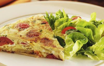 Fotografia em tons de verde em uma bancada com um prato branco redondo raso, um pano amarelo de fundo e a tortilla de omelete com batata e tomate em cima dele, ao lado, salada de alface e tomate cereja.