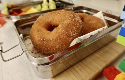 Fotografia em tons de vermelho em uma bancada de madeira clara, uma marmita ao meio com dois donuts de maçã dentro. Ao lado, brinquedos infantis.