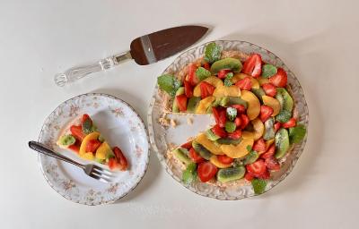 Foto vista de cima da receita de torta de frutas com creme de amêndoa, servida em um prato redondo ao lado de uma fatia cortada. A torta é decorada com frutas diversas (kiwi, manga e morango)