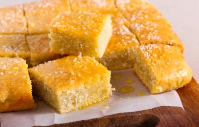 Fotografia em tons de amarelo com uma tábua ao centro. Em cima da tábua existe vários pedaços de bolo de cor amarela ao centro. Os pedaços de bolo estão cobertos com uma calda e coco ralado