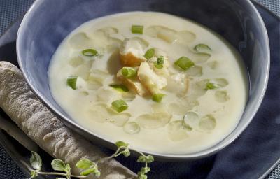 Fotografia de um recipiente azul de vidro com uma sopa de alho poró com macarrão, com cebolinha e pão por cima. O recipiente está em cima de um prato azul, sobre uma toalha de mesa da mesma cor.