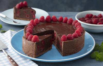 Fotografia em tons de vermelho em uma bancada de madeira azul, um pano azul quadriculado com uma espátula para bolo em cima dele. Ao centro, um prato azul redondo raso com a torta cremosa de chocolate com framboesas. Ao fundo, um prato com uma fatia.