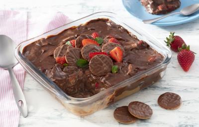 Fotografia em tons de vermelho em uma bancada de madeira clara, um pano branco com listras vermelhas, um refratário quadrado de vidro com a lasanha doce de chocolate com morangos e biscoito calipso dentro dele. Ao fundo, um prato azul com fatia do doce.