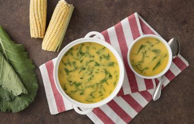 fotografia em tons de marrom, amarelo e verde de uma bancada de madeira vista de cima. Contém um pano listrado nas cores branco e rosa e por cima dois recipientes redondos com sopa de milho e ao lado duas espigas e duas folhas de couve