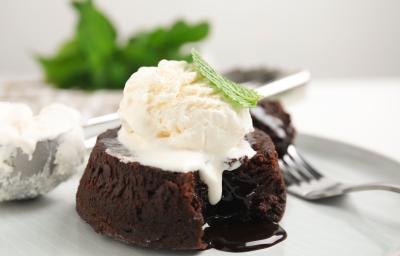 Fotografia de um petit gateau com uma bola de sorvete por cima e uma folha de hortelã. A sobremesa está em um prato raso e grande, com um garfo segurando um pedaço do bolinho e uma colher de sorvete meio cheia.