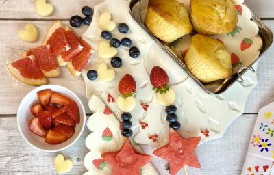 Fotografia em tons de vermelho em uma bancada de madeira clara, uma lancheira com muffin dentro, potinhos com frutas e brinquedos infantis.