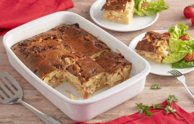 Fotografia em tons de vermelho e branco de uma travessa retangular branca com a torta sem algumas fatias, ao lado 2 pratos com fatia de torta e salada de alface e tomate. Ao fundo pano vermelho.