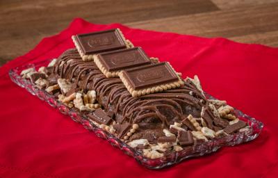 Fotografia de uma torta gelada de chocolate com chocolate derretido e alguns chocobiscuit por cima e ao lado.