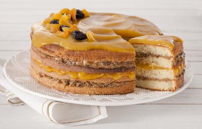 Fotografia em tons de amarelo em uma bancada de madeira branca com um pano branco com listras douradas e ao centro um prato branco com o bolo de coco com recheio de doce de leite com ameixa e geleia de damasco em cima.