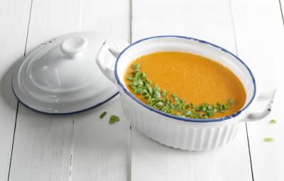 Fotografia em tons de branco, laranja e verde de uma bancada branca vista de cima. Ao centro um recipiente branco com alças e oval que contém um creme e ao lado a tampa do recipiente cor branca.