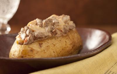 Fotografia em tons de marrom e pardo, em uma bancada contém um pano cor pardo com um garfo por cima, um recipiente marrom com uma batata com pedaços de carne com molho por cima e ao lado um copo transparente com agua