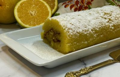 Foto da receita de Rocambole de Laranja. Observa-se um recipiente raso, retangular e branco contendo o rocambole polvilhado de açúcar de confeiteiro. Ao fundo, laranjas cortadas ao meio.