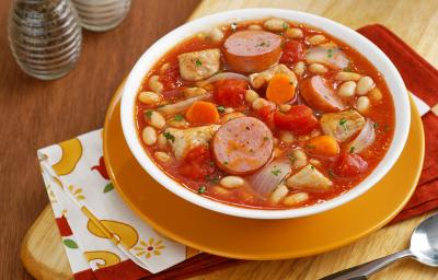 fotografia em tons de laranja e vermelho de uma bancada marrom vista de cima, contém um prato redondo laranja com um recipiente redondo branco que contém Cassoulet com pedaços de linguiça, feijão branco e abóbora.