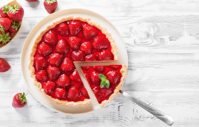 Fotografia vista de cima de um prato grande branco servindo como apoio para uma torta de morango zero lactose, com morangos por cima e uma calda brilhante. Tem um talher tirando um pedaço perfeito, e ao redor da torta tem vários morangos.