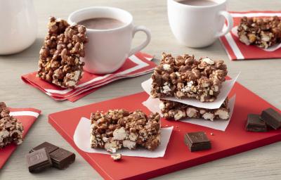 Fotografia em tons de vermelho em uma bancada de madeira clara com barras de pipoca com chocolate espalhadas pela mesa e em cima de uma tábua vermelha. Ao fundo, duas xícaras de chocolate quente com uma barra de pipoca com chocolate apoiada em uma delas.