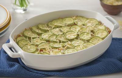 Fotografia em tons de verde em uma mesa de madeira com uma toalha branca, um pano azul, um refratário branco oval com a abobrinha gratinada dentro dele.