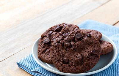 Fotografia de quatro cookies de chocolate com gotas de chocolate dentro de um recipiente de vidro branco um pouco fundo. O recipiente está por cima de um paninho azul com listras brancas sobre uma mesa de madeira.