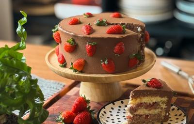Fotografia em tons de marrom com um bolo de morango ao centro. O bolo possui massa branca e é recheado com camadas de recheio de chocolate e finalizado com morangos. Fotografia em tons de marrom com um bolo de morango ao centro. O bolo possui massa branca e é recheado com camadas de recheio de chocolate e finalizado com morangos.