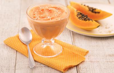 Foto em tons de laranja da receita de creme de papaia servida em uma taça de vidro sobre um pano laranja em uma mesa de madeira. Ao lado há um colher prateada e ao fundo há um prato com duas fatias médias de mamão