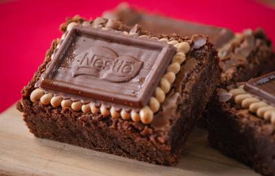 Fotografia três unidades de brownies com biscoito choco biscuit, eles estão apoiados um no outro e sobre uma tábua de madeira. O fundo da foto é vermelho.