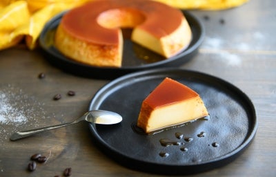 Fotografia em tons de cinza, preto, pardo e amarelo de uma bancada cinza vista de cima, Contém dois pratos redondos e pretos um contém uma fatia de pudim com uma colher para servir, e o outro o pudim sem uma fatia. Ao fundo um pano amarelo. Fotografia em tons de cinza, preto, pardo e amarelo de uma bancada cinza vista de cima, Contém dois pratos redondos e pretos um contém uma fatia de pudim com uma colher para servir, e o outro o pudim sem uma fatia. Ao fundo um pano amarelo.