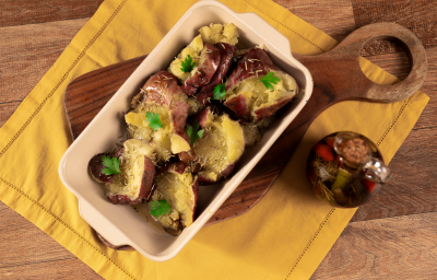 Fotografia em tons de amarelo com uma tábua de madeira ao centro. Em cima da tábua existe uma travessa clara com batatas doces ao murro gratinadas e temperadas com alecrim e azeite