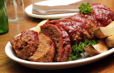 Fotografia de carne com aveia ao forno, metade cortada em fatias e dourada por cima dentro de um refratário branco oval. Ao lado, algumas batatas assadas, e ao fundo, um prato branco com um garfo encima, sobre uma mesa de madeira.