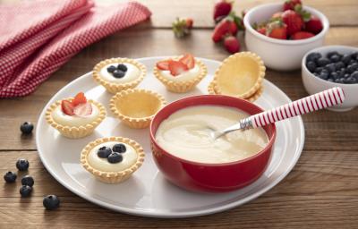 Fotografia em tons de vermelho e amarelo em uma bancada de madeira, um pano vermelho de bolinhas, um prato branco redondo com várias tortinhas recheadas com creme de confeiteiro e frutinhas. Ao lado, um potinho vermelho com o creme de confeiteiro.