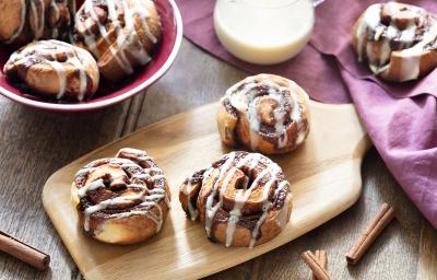 Fotografia em tons de amarelo, em uma bancada de madeira de cor marrom. Ao centro, uma tábua de madeira contendo 3 Cinnamon Rolls. Ao fundo, um pano vermelho e um recipiente contendo cinnamon rolls e uma jarra de leite.