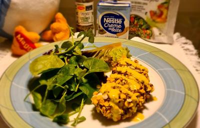 Foto aproximada de um prato redondo com bordas verde e azul com a receita de frango empanado servido com agrião. Ao fundo há produtos Maggi e Nestlé Creme de Leite e uma parte de uma pelúcia da Galinha Azul