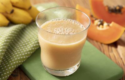 Fotografia em tons de laranja e verde de uma bancada de madeira com um suporte verde e um paninho verde com bolinhas brancas, sobre ele um copo de vidro com a vitamina de mamão e banana. Ao fundo uma fatia de mamão e um cacho de banana.