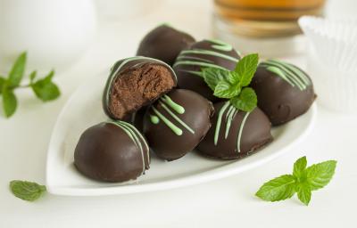 Fotografia em tons de branco, marrom e verde de uma bancada branca vista de frente, contém um prato branco com bombons de chocolate e folhas de hortelã em volta para decorar.