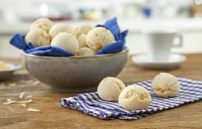 Foto de uma mesa com um pano e 3 pães de queijo redondos em cima. No fundo da foto há uma tigela com mais pães dentro