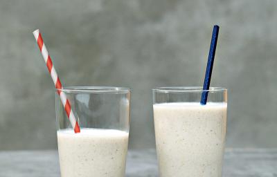 Fotografia em tons de cinza e branco de uma bancada cinza sobre ela dois copos de vidro com vitamina de banana e dois canudos, um azul e um listrado vermelho e branco.