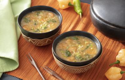 Foto em tons de bege da receita de tacacá servida em duas porções em bowls pretos com adornos em marrom sobre uma esteira de madeira. Ao fundo, há pimentas-de-cheiro e um paninho verde