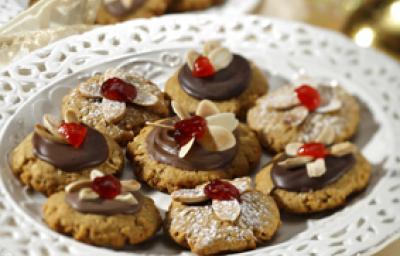 Fotografia em tons de dourado em uma mesa com uma toalha dourada, um suporte para bolo branco com detalhes em dourado e vários biscoitinhos alemães com chocolate e lascas de amêndoas.