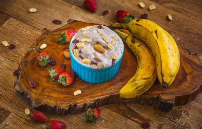 Fotografia em tons de marrom em uma bancada de madeira escura, uma tábua de tronco de árvore, um potinho azul com o sorvete de morango, banana e chia dentro dele e coberto com frutas secas. Ao lado, duas bananas e morangos espalhados pela bancada.